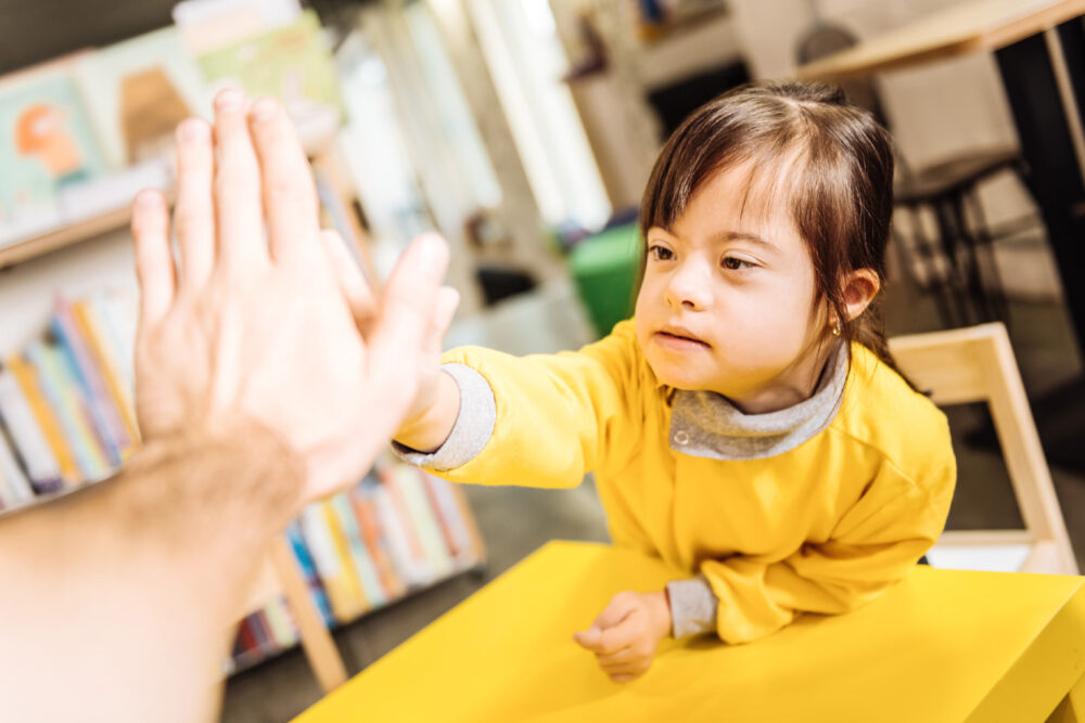 A young girl wearing a yellow shirt reaches out to give an adult a high-five while sitting at a table in a bright classroom.