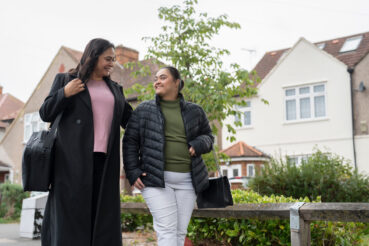 Two people walking side-by-side with houses behind them.