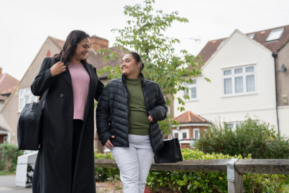 Two people walking side-by-side with houses behind them.