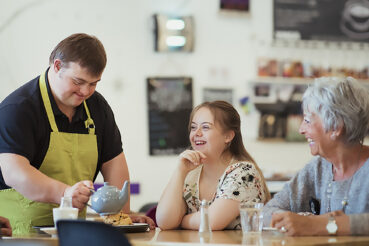 A café worker wearing a green apron pours tea for two customers sitting at a table. The customers, a younger woman and an older woman, smile and engage with him as he serves them.