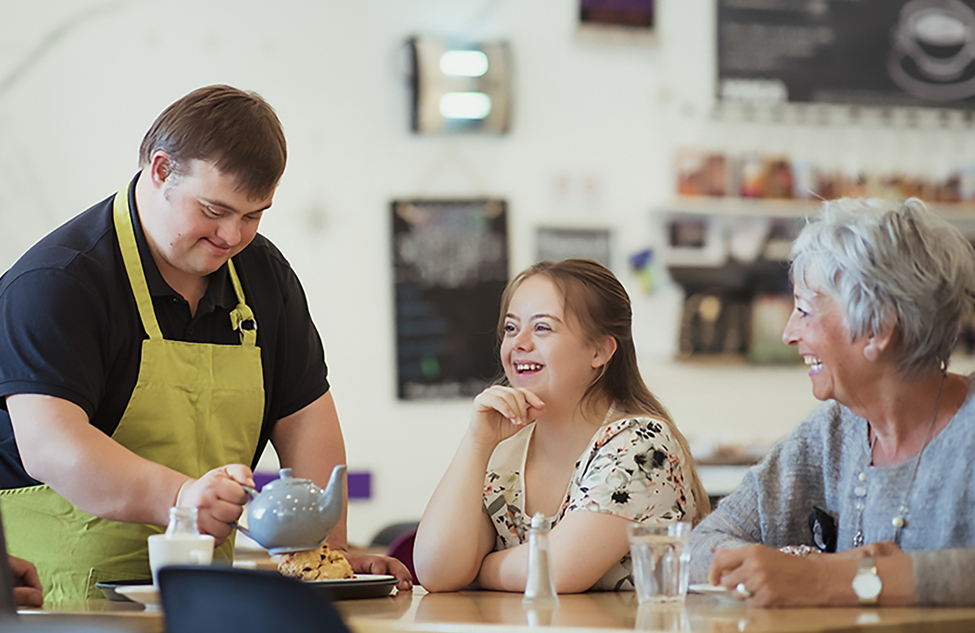 A café worker wearing a green apron pours tea for two customers sitting at a table. The customers, a younger woman and an older woman, smile and engage with him as he serves them.
