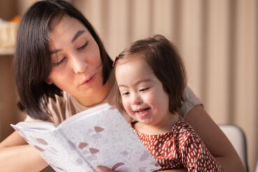 A toddler girl with special needs reads book alongside adult